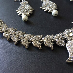 Op de afbeelding: Een zilverkleurige set bestaande uit een ketting en bijpassende oorbellen, bezet met heldere, gefacetteerde strass-steentjes in een bladvormig patroon. De oorbellen zijn aan de onderkant afgewerkt met een enkele witte kunstparel.