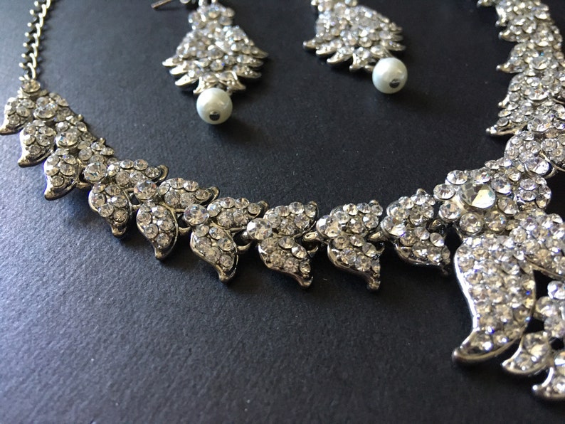 Op de afbeelding: Een zilverkleurige set bestaande uit een ketting en bijpassende oorbellen, bezet met heldere, gefacetteerde strass-steentjes in een bladvormig patroon. De oorbellen zijn aan de onderkant afgewerkt met een enkele witte kunstparel.