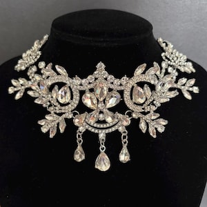 May include: A silver-toned choker necklace featuring clear, faceted crystals arranged in a floral and leaf-inspired pattern. The design includes multiple teardrop-shaped crystal dangles hanging from the centre. The text Weddingvalle is visible.