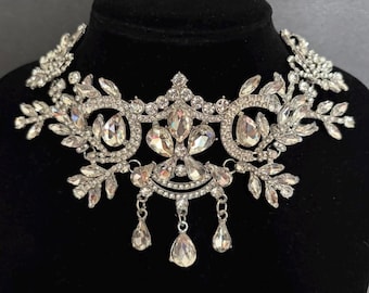 Op de afbeelding: Een zilverkleurige choker-halsketting met heldere, gefacetteerde kristallen in een bloem- en bladpatroon. Het ontwerp bevat meerdere druppelvormige kristallen hangers die in het midden hangen. De tekst Weddingvalle is zichtbaar.