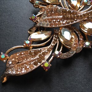 May include: Close-up of a gold-toned brooch with a floral design. The brooch features clear and amber-colored rhinestones, with iridescent accents. The leaves and stems are detailed with smaller rhinestones.