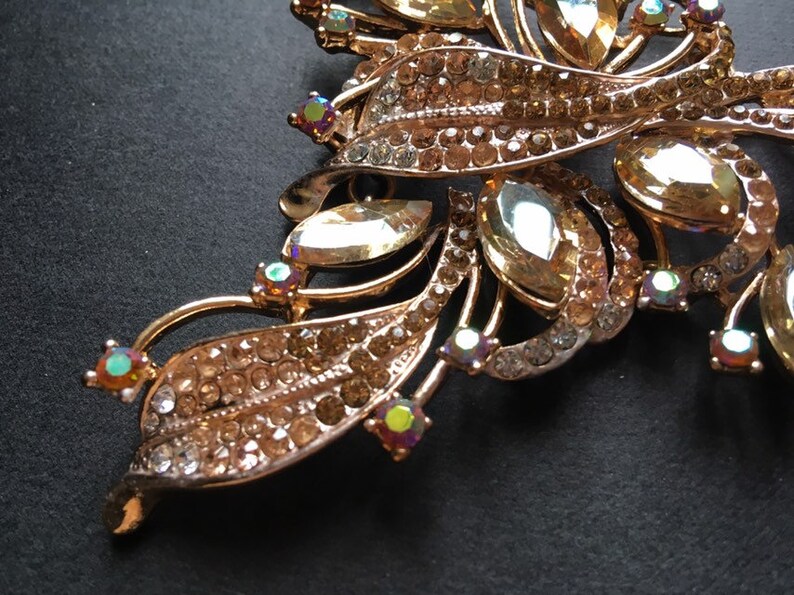 May include: Close-up of a gold-toned brooch with a floral design. The brooch features clear and amber-colored rhinestones, with iridescent accents. The leaves and stems are detailed with smaller rhinestones.