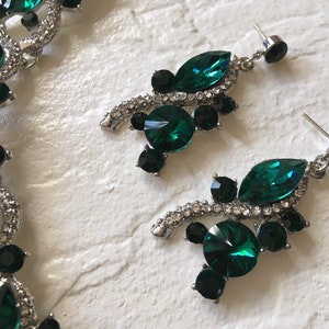 May include: A jewelry set consisting of a necklace and a pair of drop earrings. The pieces feature silver-toned metal settings adorned with faceted emerald green and black crystals, accented by small clear rhinestones along the metal edges.