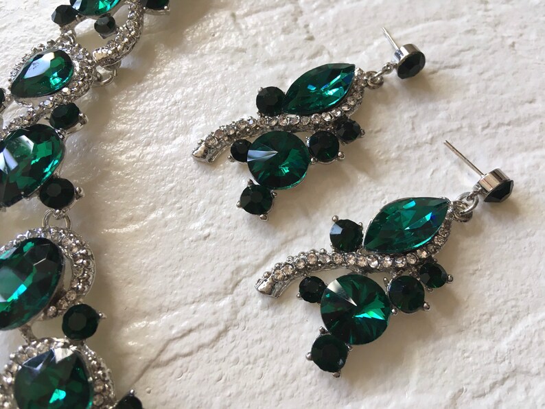 May include: A jewelry set consisting of a necklace and a pair of drop earrings. The pieces feature silver-toned metal settings adorned with faceted emerald green and black crystals, accented by small clear rhinestones along the metal edges.