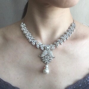 Op de afbeelding: Een zilverkleurige ketting met een reeks heldere, gefacetteerde strass-steentjes in een bladvormig patroon langs de ketting, uitlopend in een centrale decoratieve hanger die aan de onderkant een enkele, ronde, witte parel ondersteunt.