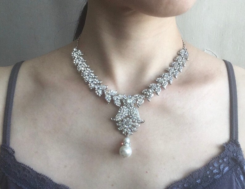 Op de afbeelding: Een zilverkleurige ketting met een reeks heldere, gefacetteerde strass-steentjes in een bladvormig patroon langs de ketting, uitlopend in een centrale decoratieve hanger die aan de onderkant een enkele, ronde, witte parel ondersteunt.