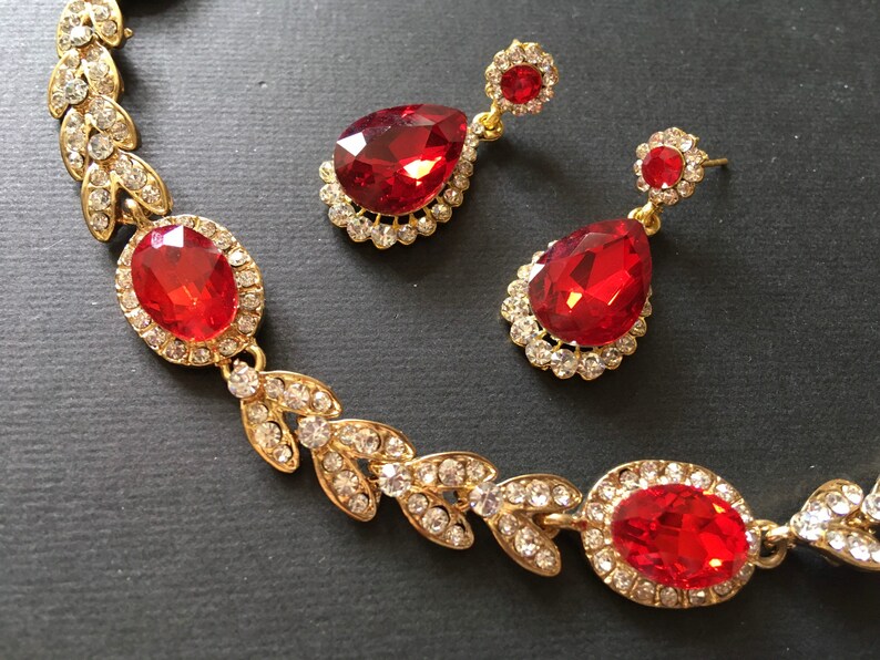 May include: A gold necklace and earrings set with red teardrop-shaped stones surrounded by clear rhinestones. The necklace features a leaf design.