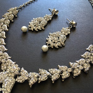 Op de afbeelding: Een zilverkleurige ketting en een bijpassend paar oorbellen met heldere kristallen strass-steentjes in bladvormige patronen. Elke oorbel en het midden van de ketting zijn voorzien van een enkele witte kunstparel die onderaan hangt.