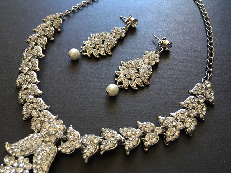 Op de afbeelding: Een zilverkleurige ketting en een bijpassend paar oorbellen met heldere kristallen strass-steentjes in bladvormige patronen. Elke oorbel en het midden van de ketting zijn voorzien van een enkele witte kunstparel die onderaan hangt.