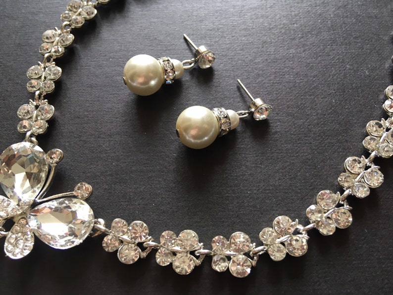 Puede incluir: Un conjunto de collar y pendientes plateados con detalles de cristal transparente y perlas de imitaci&oacute;n. El collar presenta un dise&ntilde;o de mariposa y racimos florales. Los pendientes tienen perlas colgantes y tachuelas de cristal.