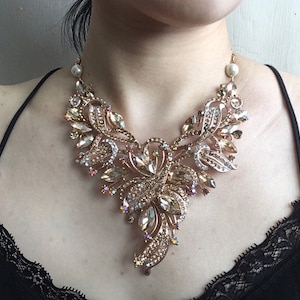May include: A gold-toned statement necklace with an elaborate floral design. The necklace features clear and champagne-colored crystals and faux pearls. The intricate design includes leaf-shaped elements and a central floral cluster, creating a glamorous look.
