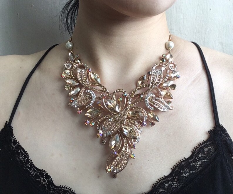 May include: A gold-toned statement necklace with an elaborate floral design. The necklace features clear and champagne-colored crystals and faux pearls. The intricate design includes leaf-shaped elements and a central floral cluster, creating a glamorous look.