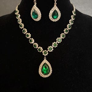 May include: A jewellery set consisting of a necklace and matching drop earrings. The necklace features a series of small green circular stones surrounded by clear crystals, with a large pear-shaped green pendant. The earrings match the pendant.