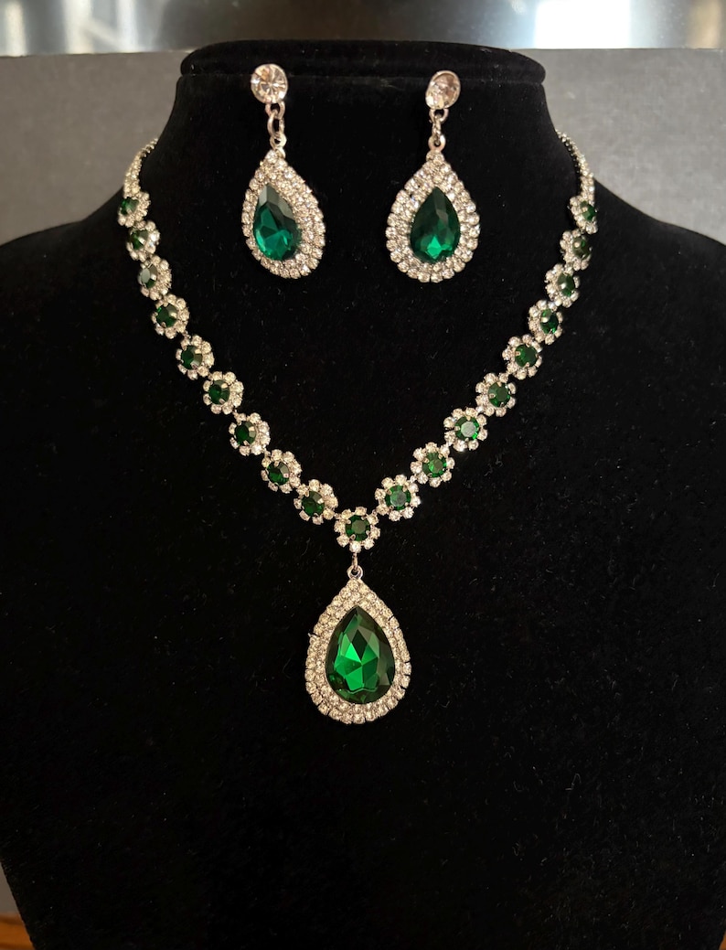 May include: A jewellery set consisting of a necklace and matching drop earrings. The necklace features a series of small green circular stones surrounded by clear crystals, with a large pear-shaped green pendant. The earrings match the pendant.