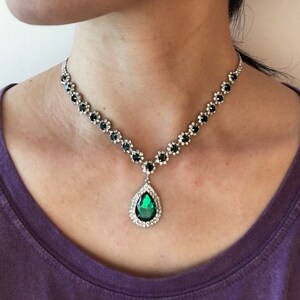 May include: A silver-toned necklace featuring a series of small, round, dark green stones set in metallic flower-shaped frames, leading to a central teardrop-shaped pendant with a large, faceted emerald-green stone surrounded by clear crystals.