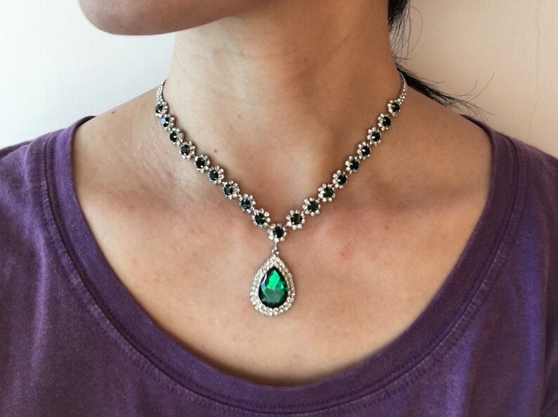May include: A silver-toned necklace featuring a series of small, round, dark green stones set in metallic flower-shaped frames, leading to a central teardrop-shaped pendant with a large, faceted emerald-green stone surrounded by clear crystals.