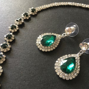 May include: A jewellery set consisting of a necklace and two drop earrings. The necklace features a silver-tone chain with circular links containing dark green stones surrounded by clear crystals. The earrings have a teardrop-shaped green stone.