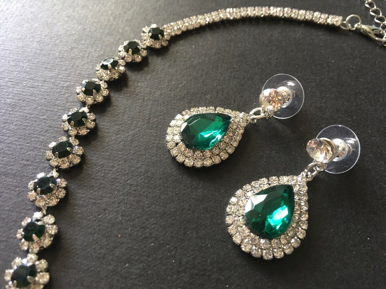 May include: A jewellery set consisting of a necklace and two drop earrings. The necklace features a silver-tone chain with circular links containing dark green stones surrounded by clear crystals. The earrings have a teardrop-shaped green stone.