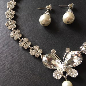 Puede incluir: Un conjunto de joyas plateadas con un collar y pendientes a juego. El collar tiene un colgante de mariposa con cristales transparentes y brillantes y una perla colgante. Los pendientes tienen perlas.