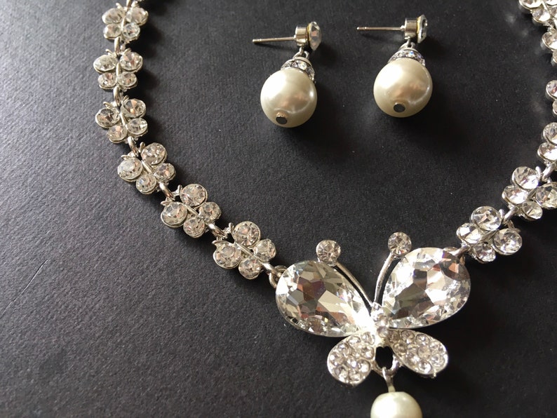 Puede incluir: Un conjunto de joyas plateadas con un collar y pendientes a juego. El collar tiene un colgante de mariposa con cristales transparentes y brillantes y una perla colgante. Los pendientes tienen perlas.