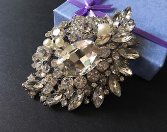 May include: A large, ornate brooch with a silver-tone metal setting. The brooch is encrusted with numerous clear, faceted crystals of various shapes and sizes, including a large oval center stone and several faux pearls. A light blue gift box with a purple ribbon is in the background.