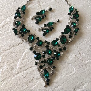 May include: A silver-toned necklace and earring set featuring large emerald green teardrop and round crystals, accented with smaller black stones and clear pav&eacute; rhinestones arranged in a swirling, ornate metal design.