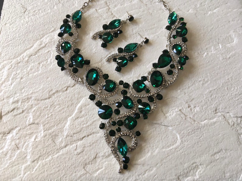 May include: A silver-toned necklace and earring set featuring large emerald green teardrop and round crystals, accented with smaller black stones and clear pav&eacute; rhinestones arranged in a swirling, ornate metal design.