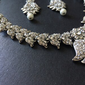 Op de afbeelding: Een zilverkleurige ketting en bijpassende oorbellen met ingewikkelde bladvormige schakels ingelegd met heldere, sprankelende strass-steentjes. De oorbellen zijn voorzien van een witte parel aan de onderkant van elk bladvormig ontwerp.