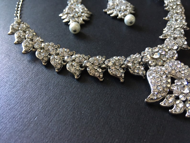 Op de afbeelding: Een zilverkleurige ketting en bijpassende oorbellen met ingewikkelde bladvormige schakels ingelegd met heldere, sprankelende strass-steentjes. De oorbellen zijn voorzien van een witte parel aan de onderkant van elk bladvormig ontwerp.