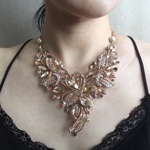 May include: A statement necklace with a gold-tone metal base, adorned with numerous clear and amber-colored crystals. The necklace features an intricate floral design with a pearl accent near the clasp. The necklace is worn on a person wearing a black top.