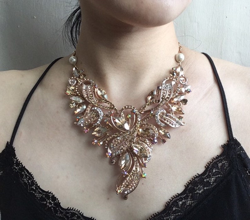 May include: A statement necklace with a gold-tone metal base, adorned with numerous clear and amber-colored crystals. The necklace features an intricate floral design with a pearl accent near the clasp. The necklace is worn on a person wearing a black top.