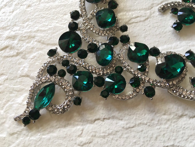 May include: A silver-toned necklace featuring large emerald green faceted crystals, smaller round black stones, and clear pav&eacute; rhinestones set in a swirling metal frame. The design incorporates various shapes including marquise and round cuts.