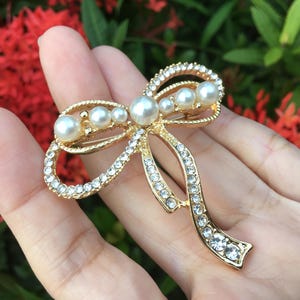 May include: A gold-toned bow brooch with faux pearls and clear rhinestones. The bow features a central knot with a cluster of pearls and rhinestone-lined loops and tails. The brooch is set against a blurred background of red and green foliage.