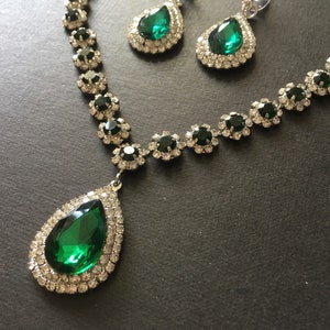 May include: A jewellery set consisting of a necklace and matching earrings. The necklace features a large teardrop-shaped emerald green stone pendant surrounded by clear crystals, with a chain of smaller green stones and clear crystal accents.