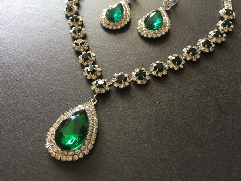 May include: A jewellery set consisting of a necklace and matching earrings. The necklace features a large teardrop-shaped emerald green stone pendant surrounded by clear crystals, with a chain of smaller green stones and clear crystal accents.