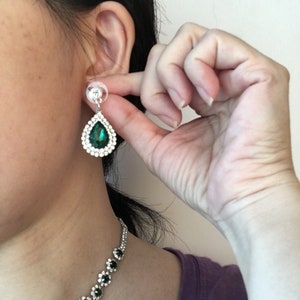 May include: A teardrop-shaped earring featuring a central dark green gemstone surrounded by a halo of small clear crystals set in a silver-toned metal frame, accompanied by a matching necklace with a series of green and clear crystal floral links.