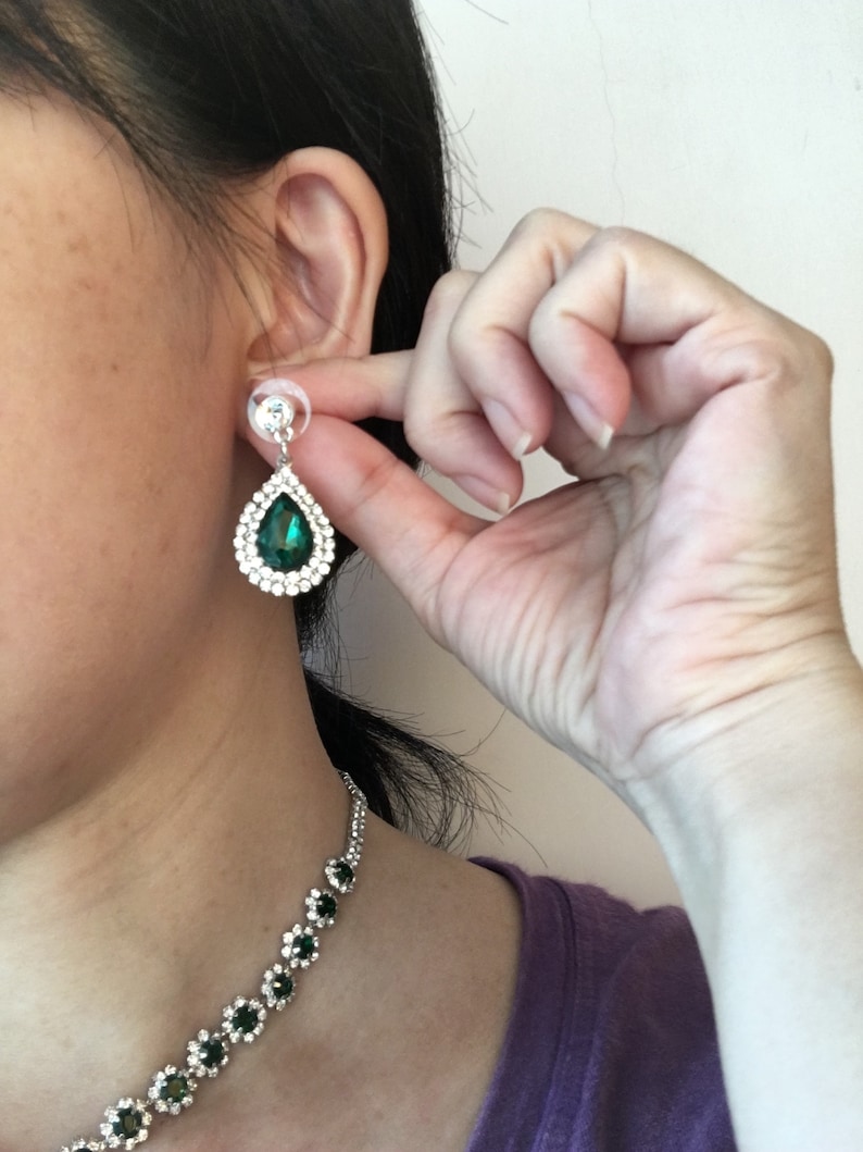 May include: A teardrop-shaped earring featuring a central dark green gemstone surrounded by a halo of small clear crystals set in a silver-toned metal frame, accompanied by a matching necklace with a series of green and clear crystal floral links.