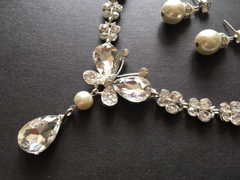 Puede incluir: Un conjunto de joyas plateadas que incluye un collar y pendientes a juego. El collar presenta un colgante en forma de l&aacute;grima y un dise&ntilde;o de mariposa con cristales transparentes y brillantes. Los pendientes son de tipo perla.