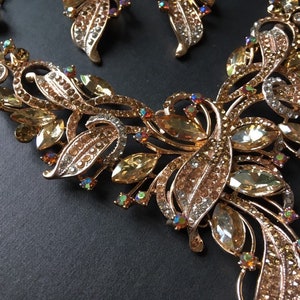 May include: A gold-toned jewelry set featuring a necklace and matching earrings. The necklace has a floral design with amber-colored and clear crystals. The earrings mirror the necklace's design. The set is on a dark background.