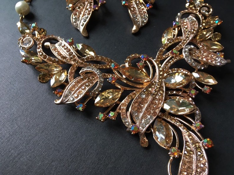 May include: A gold-toned jewelry set featuring a necklace and matching earrings. The necklace has a floral design with amber-colored and clear crystals. The earrings mirror the necklace's design. The set is on a dark background.