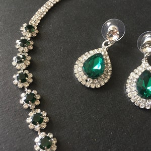 May include: A jewellery set consisting of a necklace and two drop earrings. The necklace features a series of circular links with dark green stones surrounded by clear crystals. The earrings have pear-shaped dark green stones with clear crystal halos.