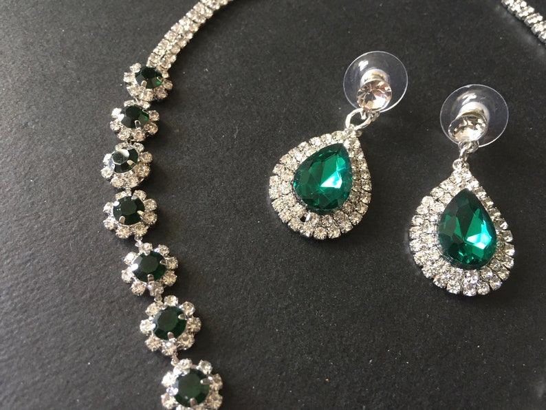 May include: A jewellery set consisting of a necklace and two drop earrings. The necklace features a series of circular links with dark green stones surrounded by clear crystals. The earrings have pear-shaped dark green stones with clear crystal halos.