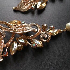 May include: Close-up of a gold-toned necklace with leaf and floral designs. The necklace features clear and amber-colored rhinestones, and a single pearl. The intricate details and sparkling accents create an elegant aesthetic.