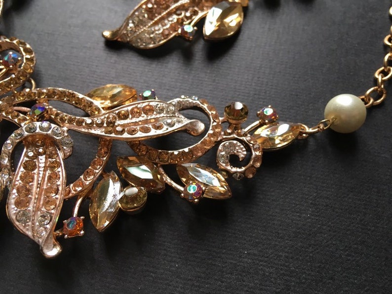 May include: Close-up of a gold-toned necklace with leaf and floral designs. The necklace features clear and amber-colored rhinestones, and a single pearl. The intricate details and sparkling accents create an elegant aesthetic.