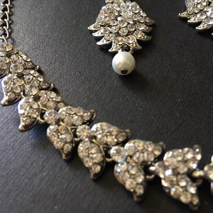 Op de afbeelding: Een zilverkleurige ketting- en oorbellenset met bladvormige schakels ingelegd met heldere strass-steentjes. Elk sieraad is voorzien van een enkele witte kunstparel. De ketting heeft een metalen schakelketting voor een verstelbare lengte.