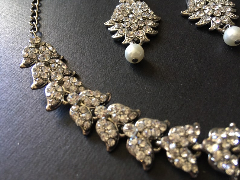 Op de afbeelding: Een zilverkleurige ketting- en oorbellenset met bladvormige schakels ingelegd met heldere strass-steentjes. Elk sieraad is voorzien van een enkele witte kunstparel. De ketting heeft een metalen schakelketting voor een verstelbare lengte.