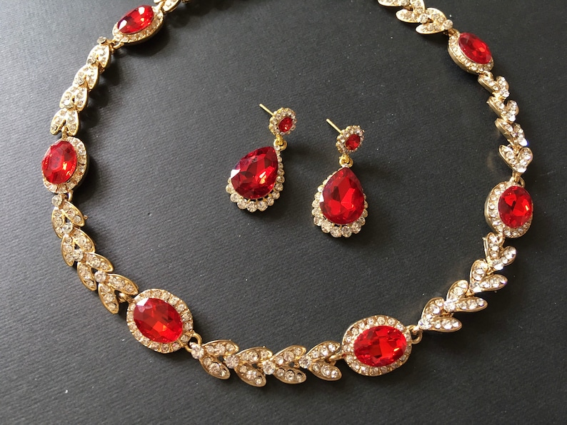 May include: A gold necklace with red crystal stones and matching earrings. The necklace features a leaf design with small crystals.