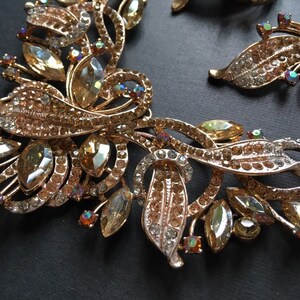 May include: A gold-toned jewelry set featuring a necklace and earrings. The pieces are adorned with clear and amber-colored crystals and rhinestones, arranged in a floral and leaf-like design. The jewelry is set against a black background.