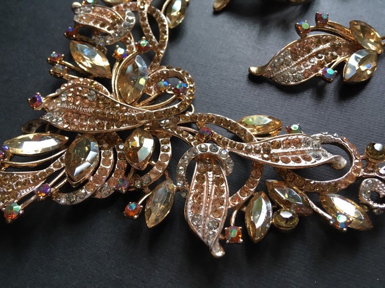 May include: A gold-toned jewelry set featuring a necklace and earrings. The pieces are adorned with clear and amber-colored crystals and rhinestones, arranged in a floral and leaf-like design. The jewelry is set against a black background.