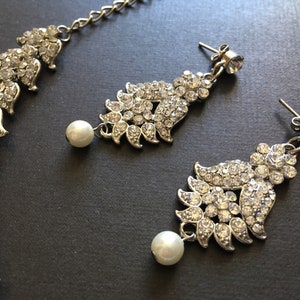 Op de afbeelding: Een zilverkleurige sieradenset bestaande uit een ketting en twee oorbellen. Elk sieraad heeft een bladvormig ontwerp ingelegd met heldere strass-steentjes en een enkele witte kunstparel die aan de onderkant van de oorbelhangers bungelt.